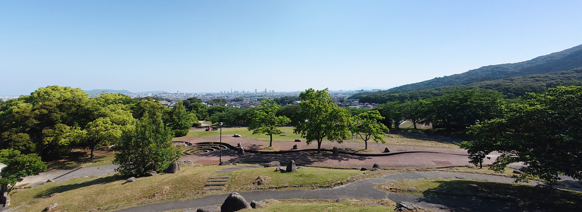 西油山中央公園（シンデレラ公園）の高台から見渡す福岡市の全景。手前に緑豊かな木々や芝生の広場、奥に福岡タワー方面の街並みと広大な青空が広がる景観。