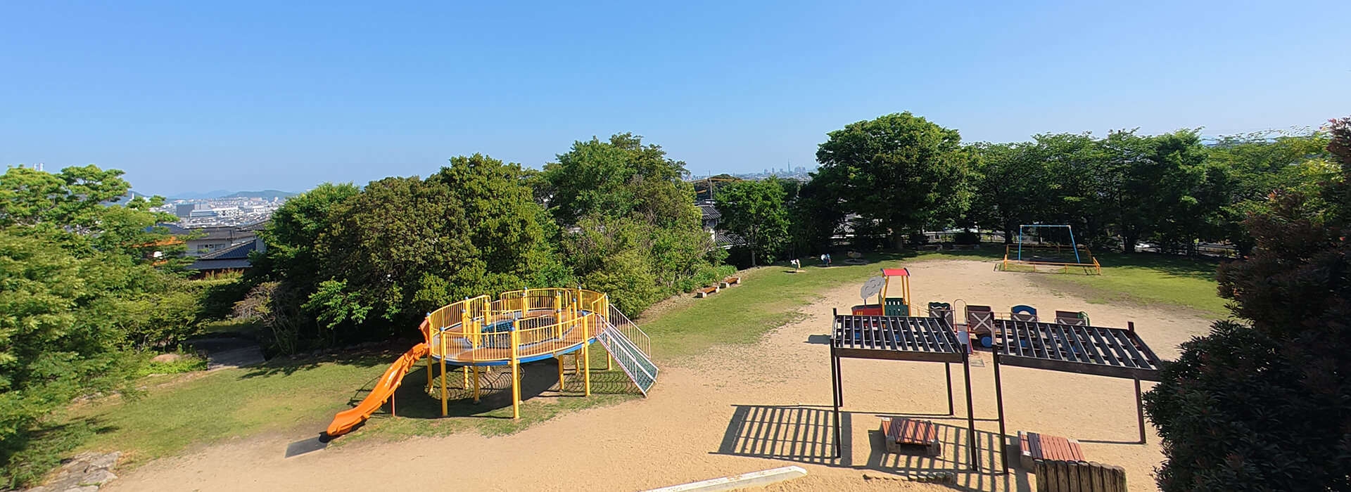 西油山中央公園の遊具エリア。黄色いネット遊具や汽車の形をした幼児用遊具、ブランコ、砂場、手前にある休憩スペースの骨組みが芝生広場に並ぶ風景。