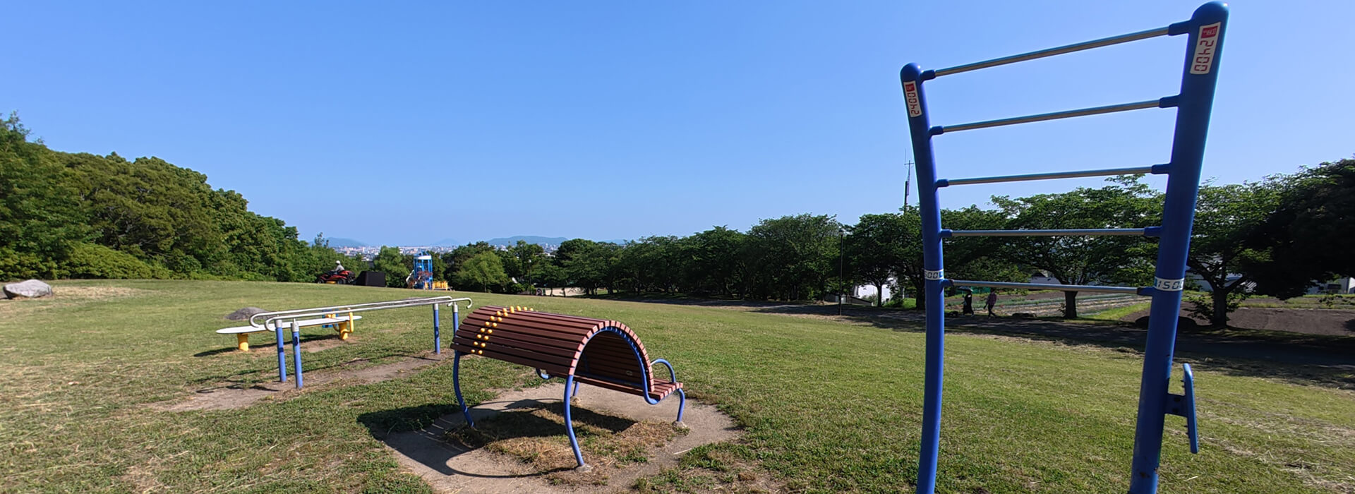 西油山中央公園の芝生スペースに設置された健康遊具。手前に青いストレッチ用の器具や茶色の背伸ばしベンチ、奥に平行棒が並ぶなだらかな斜面の広場。