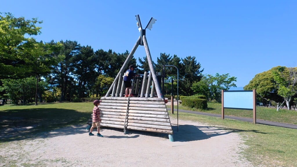 今津運動公園のアスレチック広場にある「タワーアドベンチャー」。丸太を組んで作られた三角形のタワーを登って遊ぶ子供たちの姿。