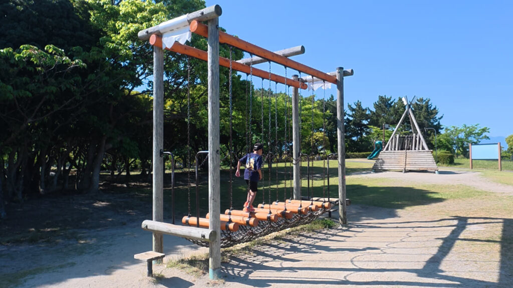 今津運動公園のアスレチック広場にある「ステップアドベンチャー」。チェーンで吊るされた丸太の上をバランスを取りながら進む子供の姿。