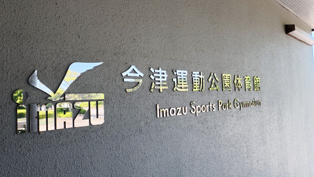 今津運動公園体育館の入り口壁面にある、シルバーに輝くスタイリッシュな施設名看板とロゴマーク。