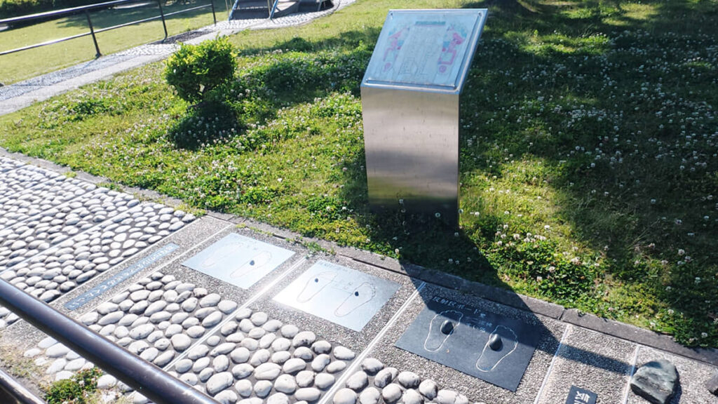 今津運動公園のリフレッシュロード脇に立つ金属製の足裏ツボ解説板。手前の路面には、乗ると特定のツボを刺激できる3つの黒い足型パネルが埋め込まれている。
