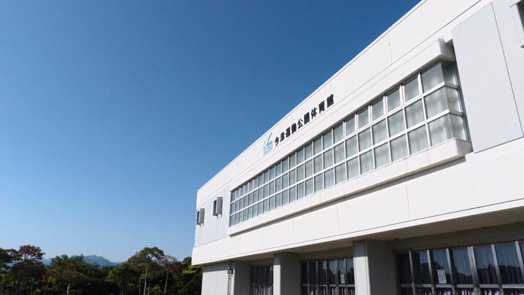 青空に映える今津運動公園体育館の白い外観建物。壁面には「今津運動公園体育館」の文字が記されている。