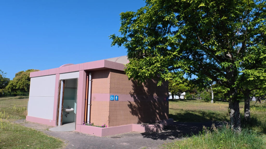 今津運動公園の球技場（芝生）B（第6駐車場側）にある公衆トイレ外観。ベージュの壁にピンク色のラインが入った四角い建物。