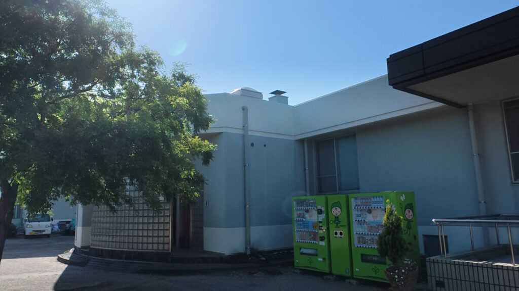 今津運動公園の管理棟（体育館）の入り口横にあるトイレエリア。手前にはカエルのキャラクターが描かれた緑色の自動販売機が並んでいる。