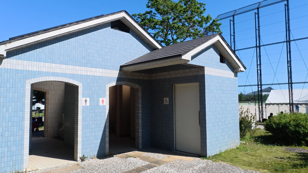 今津運動公園の第5駐車場と野球場・球技場の間にある青いタイル調の公衆トイレ外観。奥に野球場の防球ネットが見える。