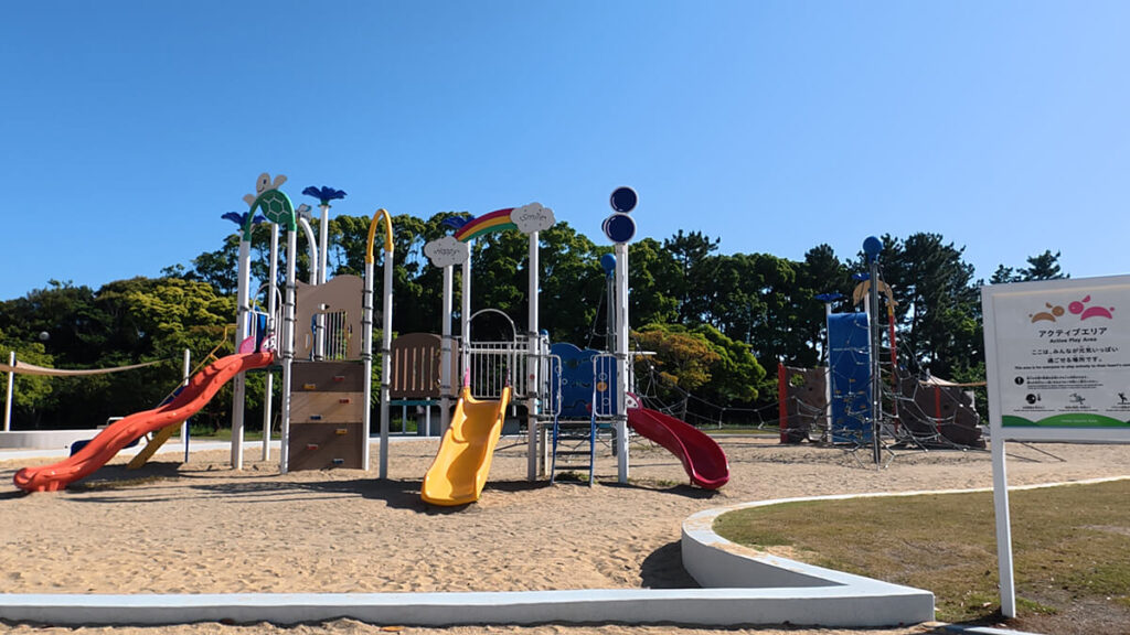 今津運動公園のアクティブエリアにある大型複合遊具の全景。砂場にカラフルなすべり台やボルダリング、手前に白い案内看板が立っている。