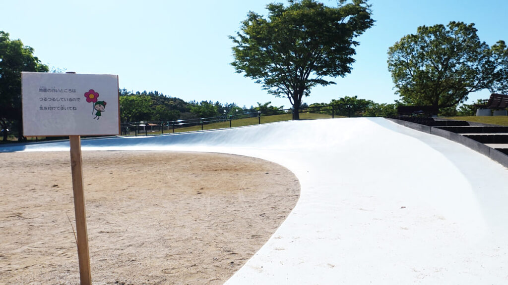 今津運動公園の白いなだらかな傾斜遊具。「地面の白いところはつるつるしているよ」と書かれた案内看板。