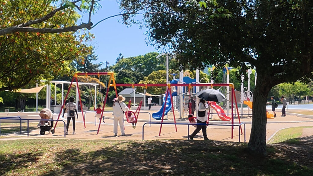今津運動公園の遊具広場にある赤と黄色のブランコ。カゴ型の安全なブランコで遊ぶ親子や、奥に見える青と赤の複合すべり台、周囲の木々の風景。