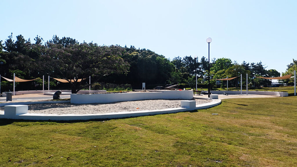 今津運動公園の「みんなの広場」に新設されたインクルーシブ遊具エリアの全景 。白い囲いのある砂場や日よけのタープ、周囲の青空と芝生の風景。