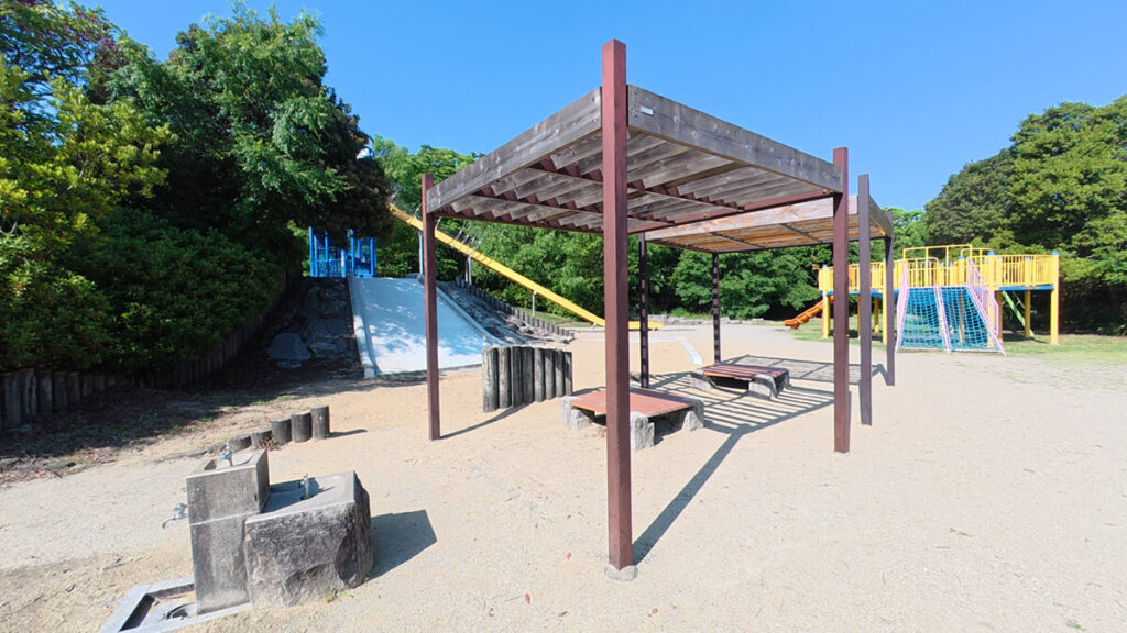 西油山中央公園の遊具広場横にある休憩スペース 。木製のベンチが並び、手前には日よけの骨組みが立っている 。