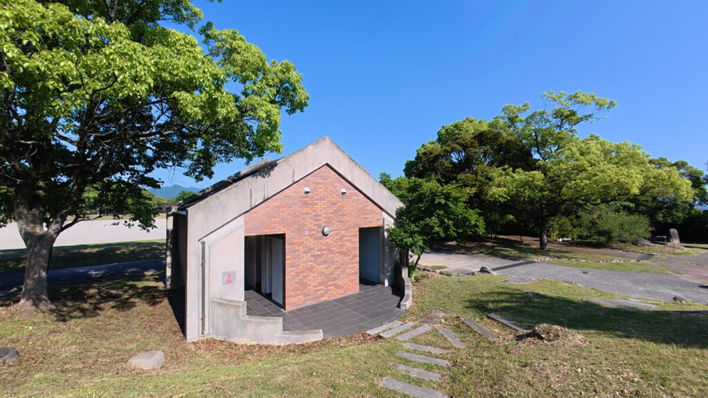 西油山中央公園のグラウンド（球技場）そばにあるベージュ色の公衆トイレ。緑の樹木に囲まれた平屋の建物。