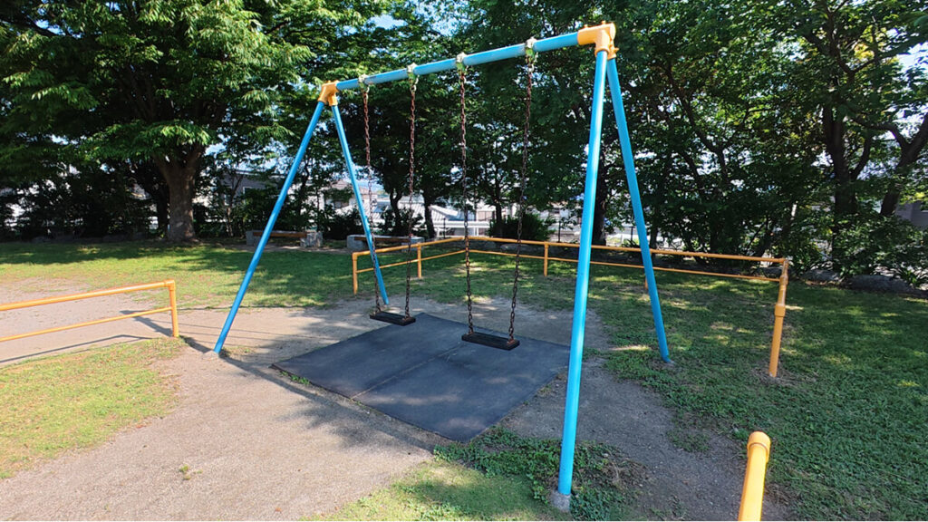 西油山中央公園のちびっこ広場にある、青いフレームのシンプルな二人乗りブランコと砂地の地面 。